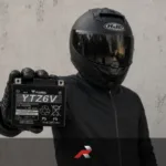 Yuasa motorcycle battery held by a rider in full motorcycle gear with a full-face helmet against a textured wall