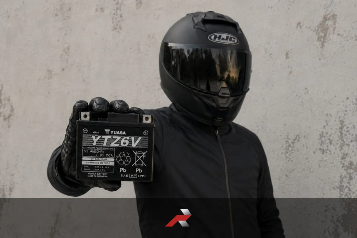 Yuasa motorcycle battery held by a rider in full motorcycle gear with a full-face helmet against a textured wall